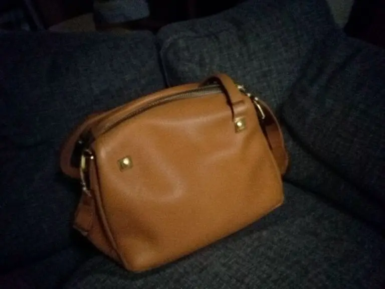 Tan leather handbag with gold studs lying tilted on a dark gray sofa in dim light