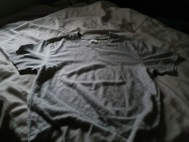 Wrinkled heather gray crew neck t-shirt lying flat on an unmade bed in dim light